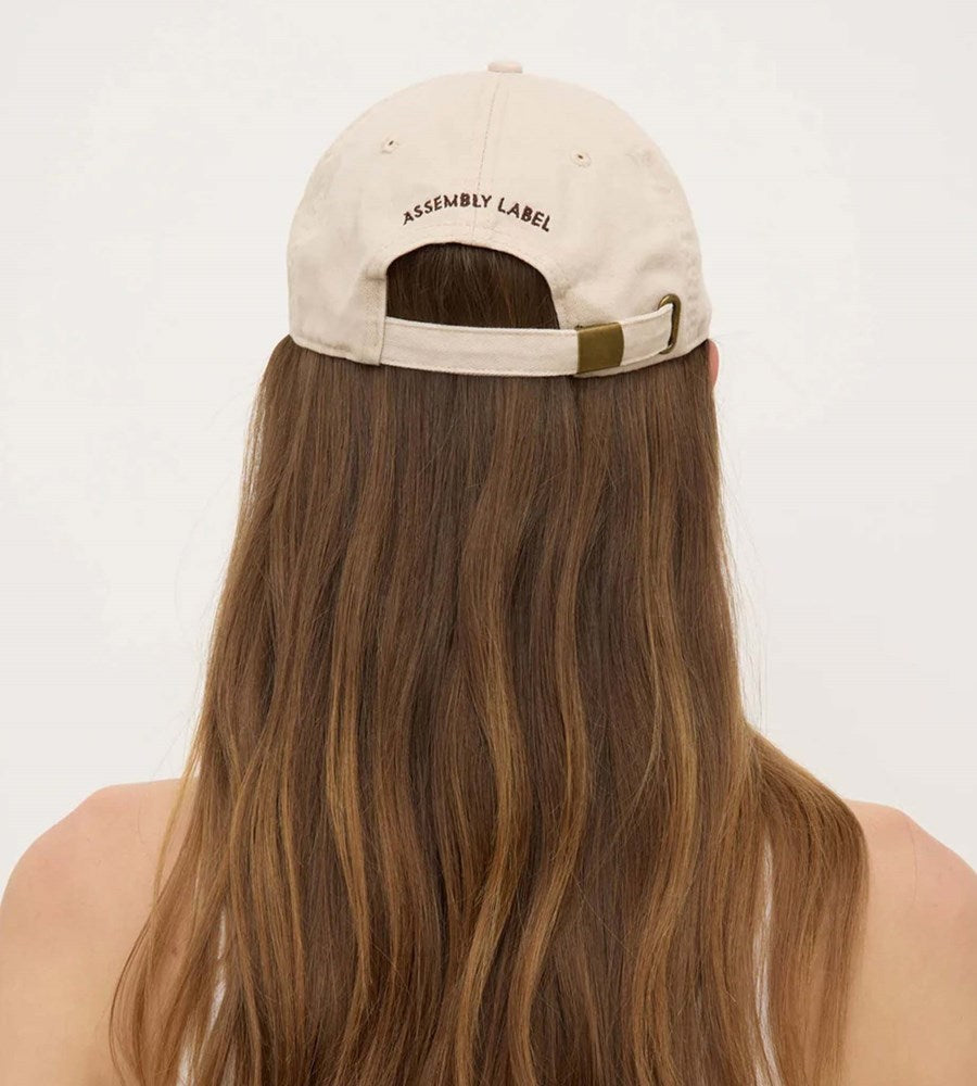 Beige cap with 'Assembly Label' on the back worn by a person with long hair, against a white background
