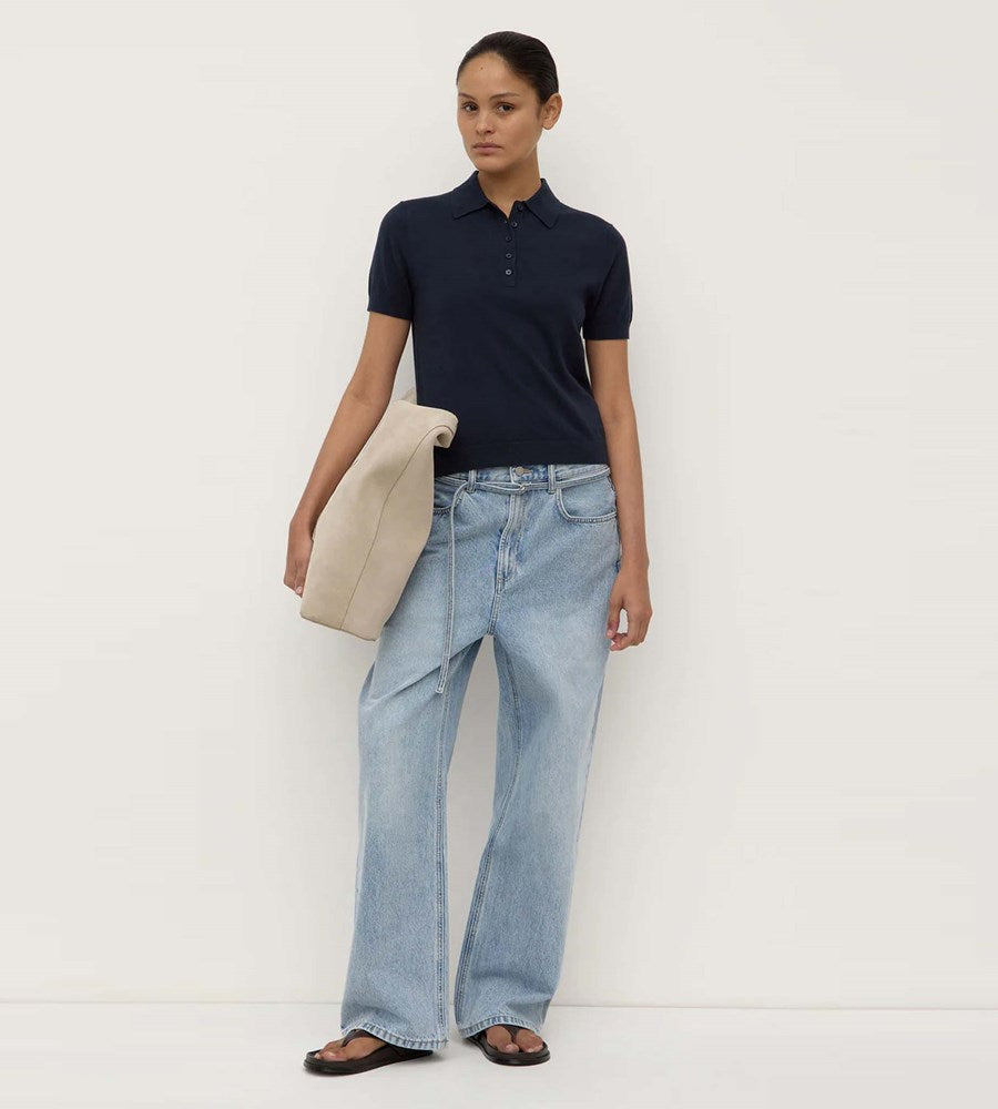 Person wearing a navy polo shirt and light blue jeans holding a beige clutch against a white background