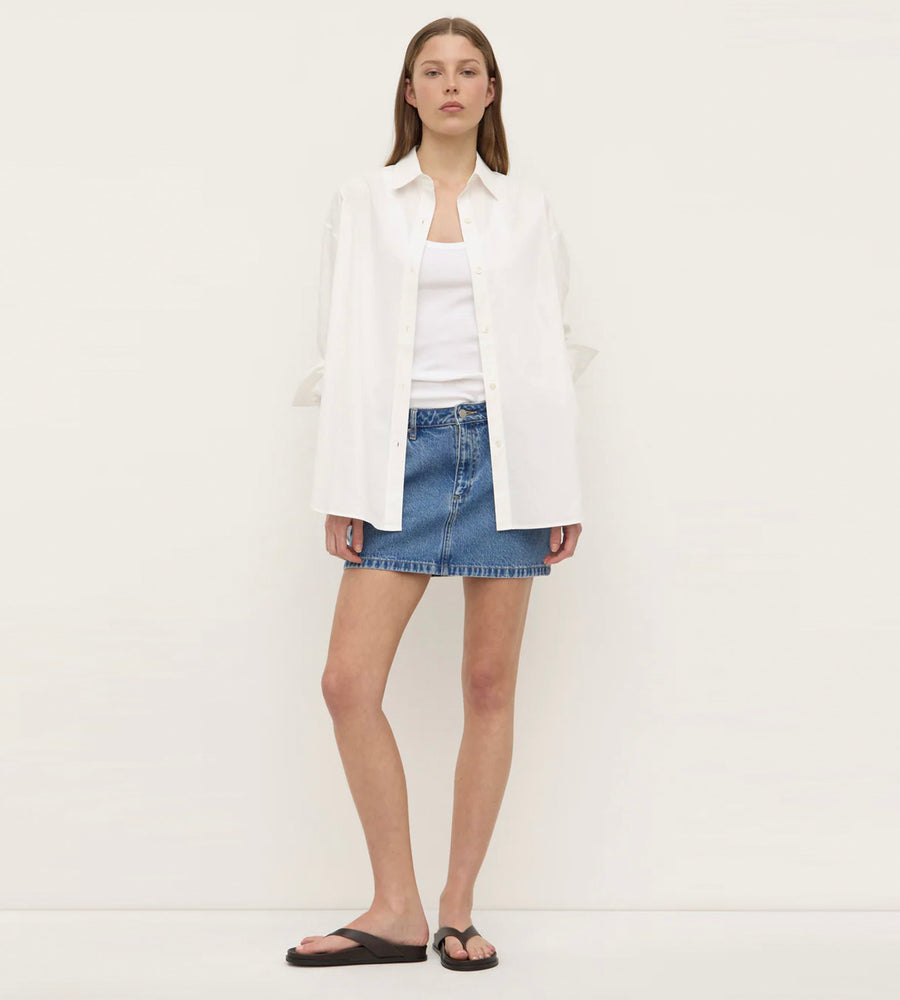 Woman wearing a white blazer, white shirt, and denim skirt on a plain background