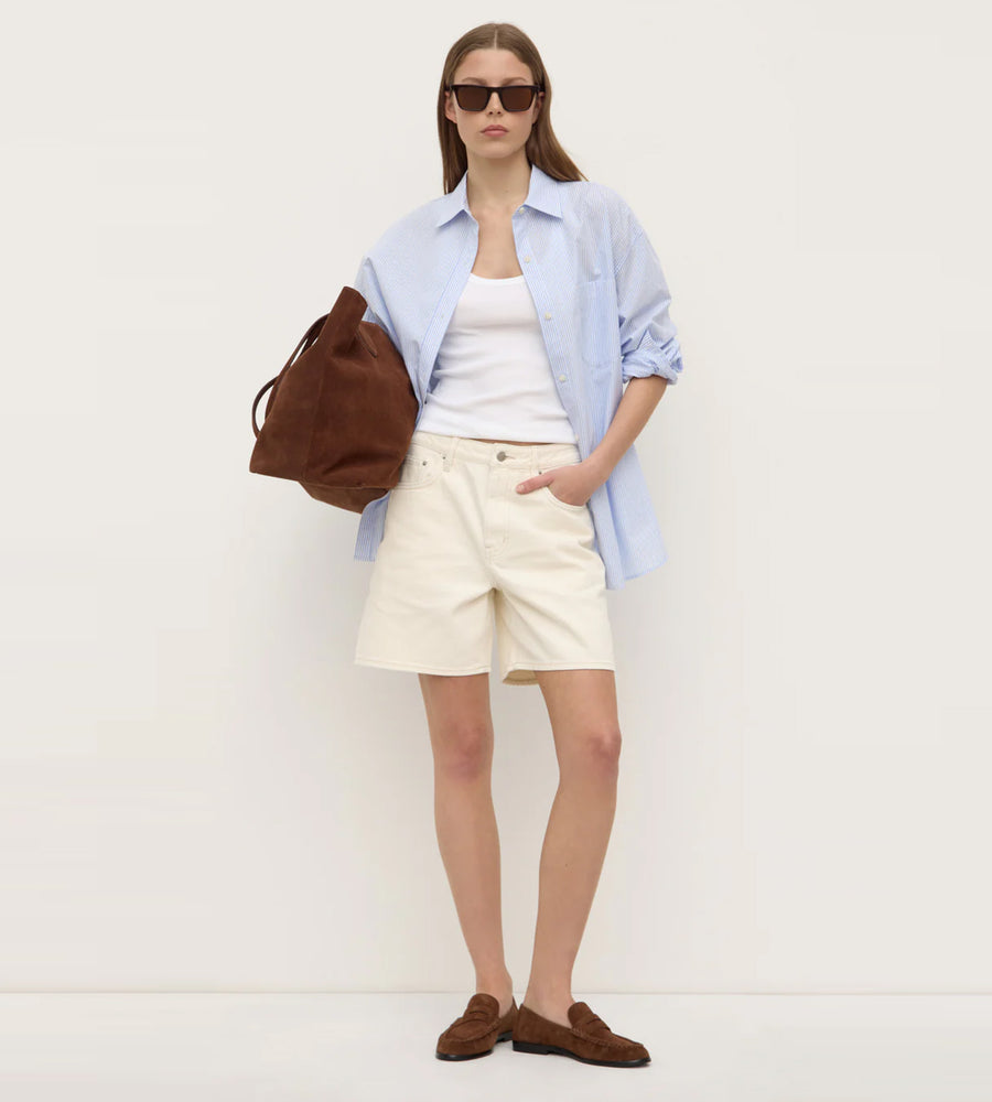 Woman wearing a light blue shirt, white shorts, and brown loafers with a brown bag on a white background