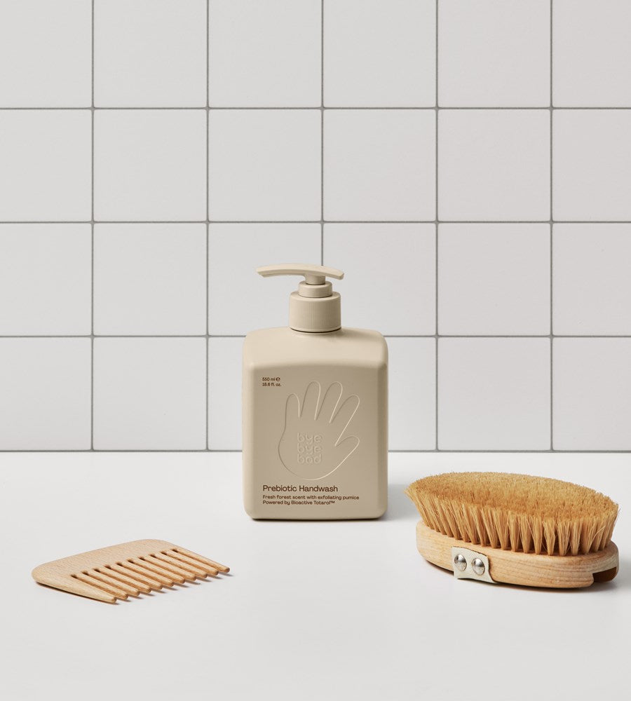 Beige hand soap bottle with pump, wooden scrubber, and soap dish on a tiled bathroom floor.