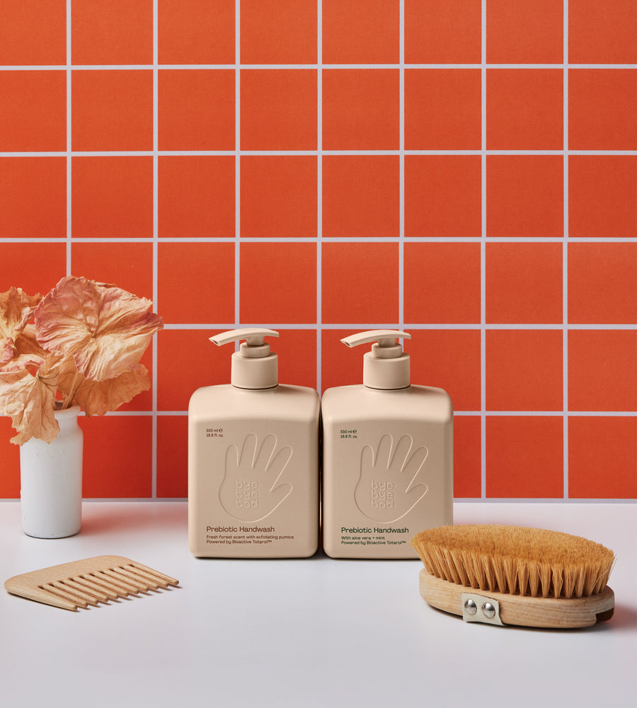 Two beige hand soap bottles with a scrubber and a vase against an orange tiled wall.