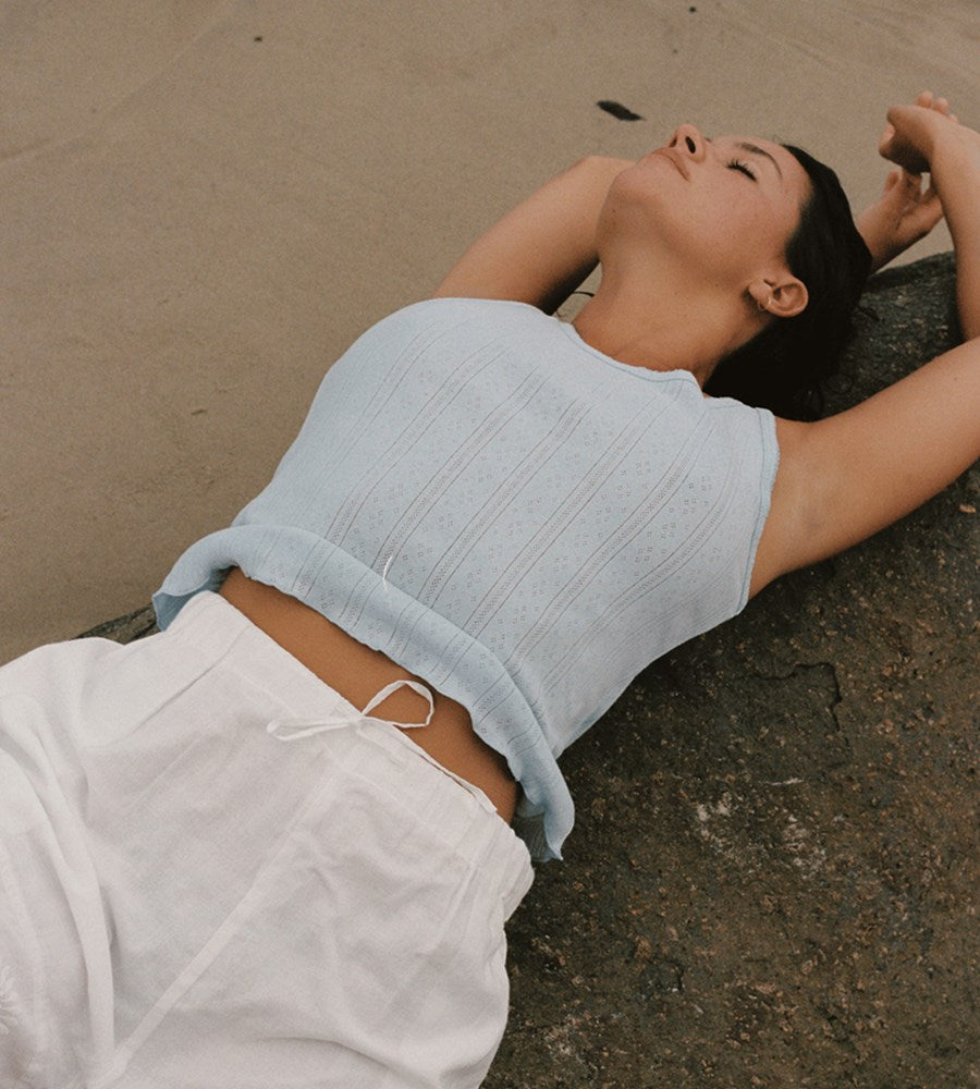 Person lying on a sandy surface wearing a light blue top and white pants.