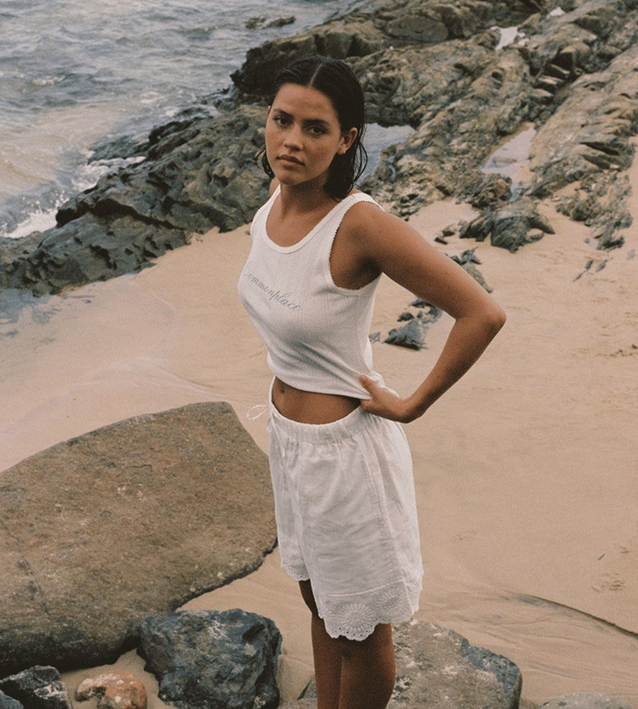 Woman in white tank top and skirt standing on a rocky beach.