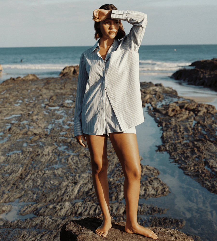 Woman standing on a rocky beach wearing a light blue shirt and shorts.