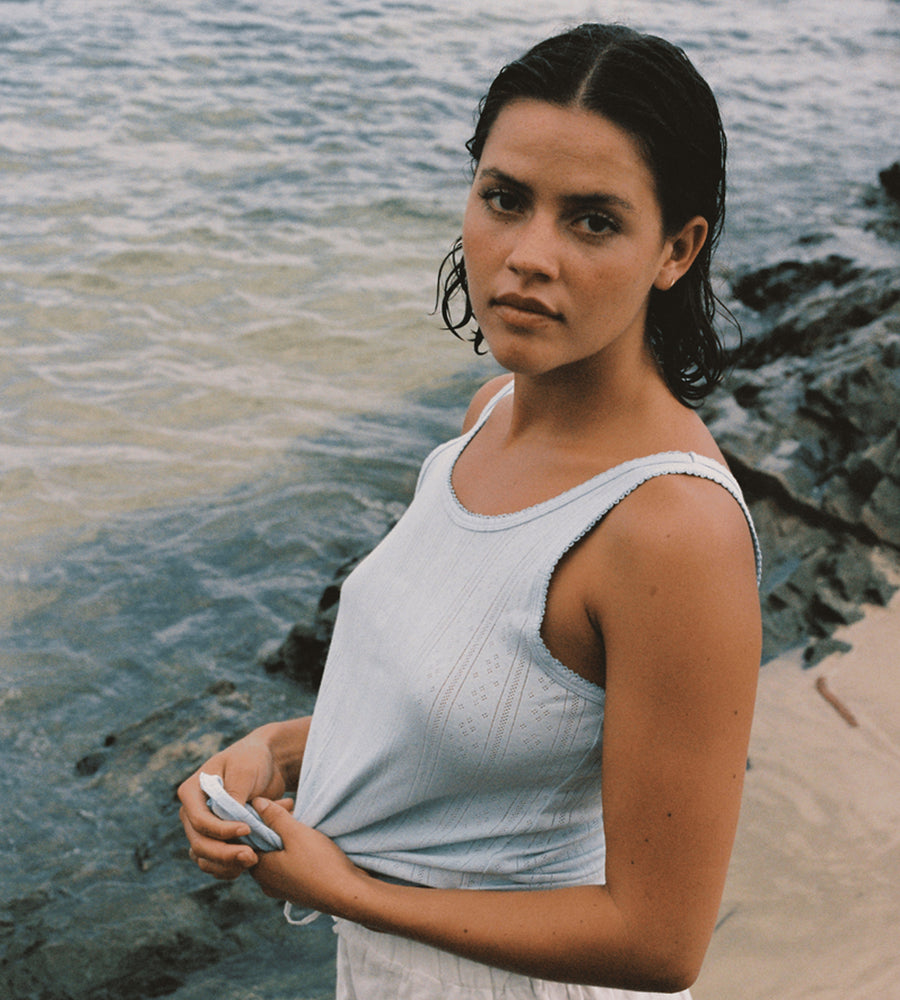 Woman standing by the water wearing a light blue tank top.