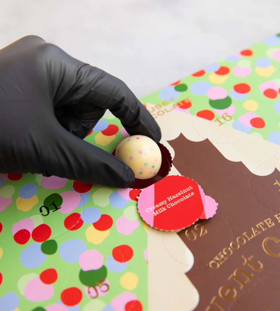 Hand wearing a black glove holding a chocolate truffle over a colorful packaging with a red label.