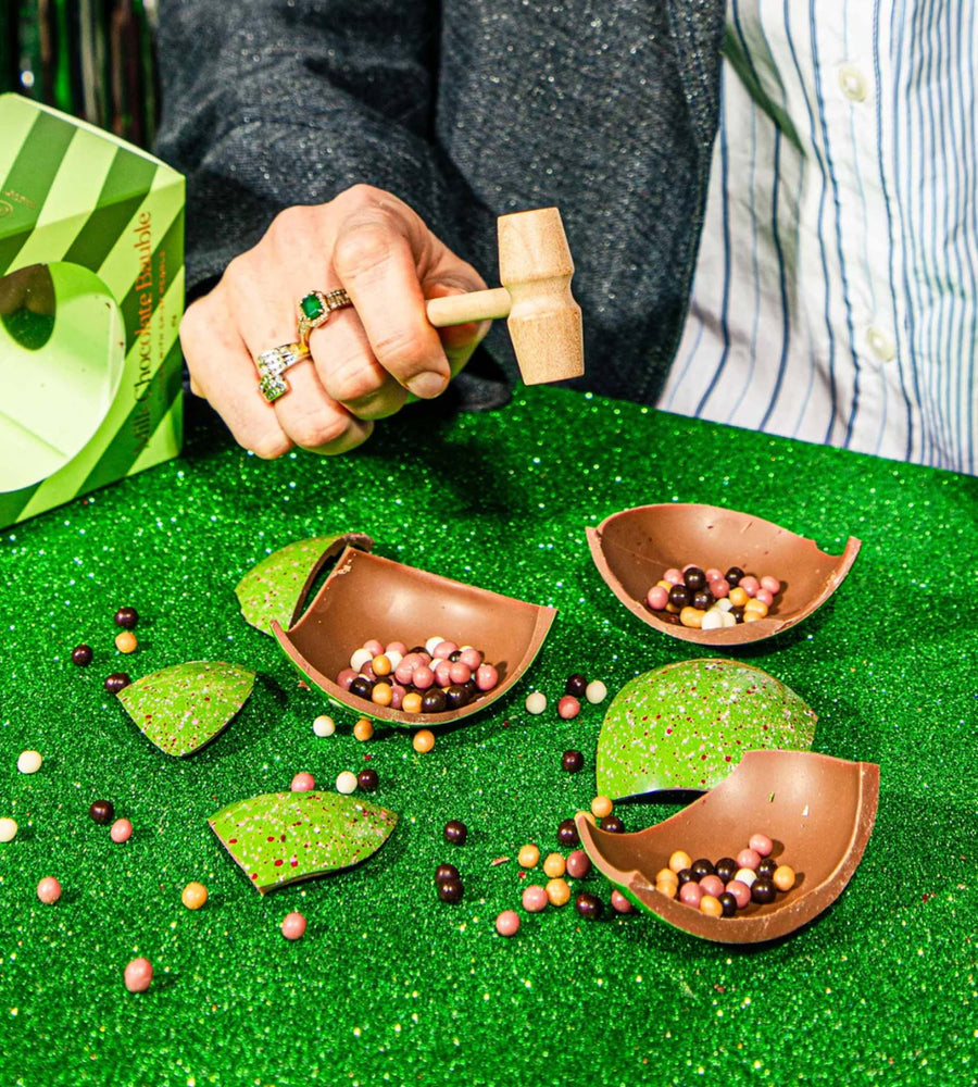 Person using a wooden mallet to fill small brown bowls with colorful beads on a green surface.