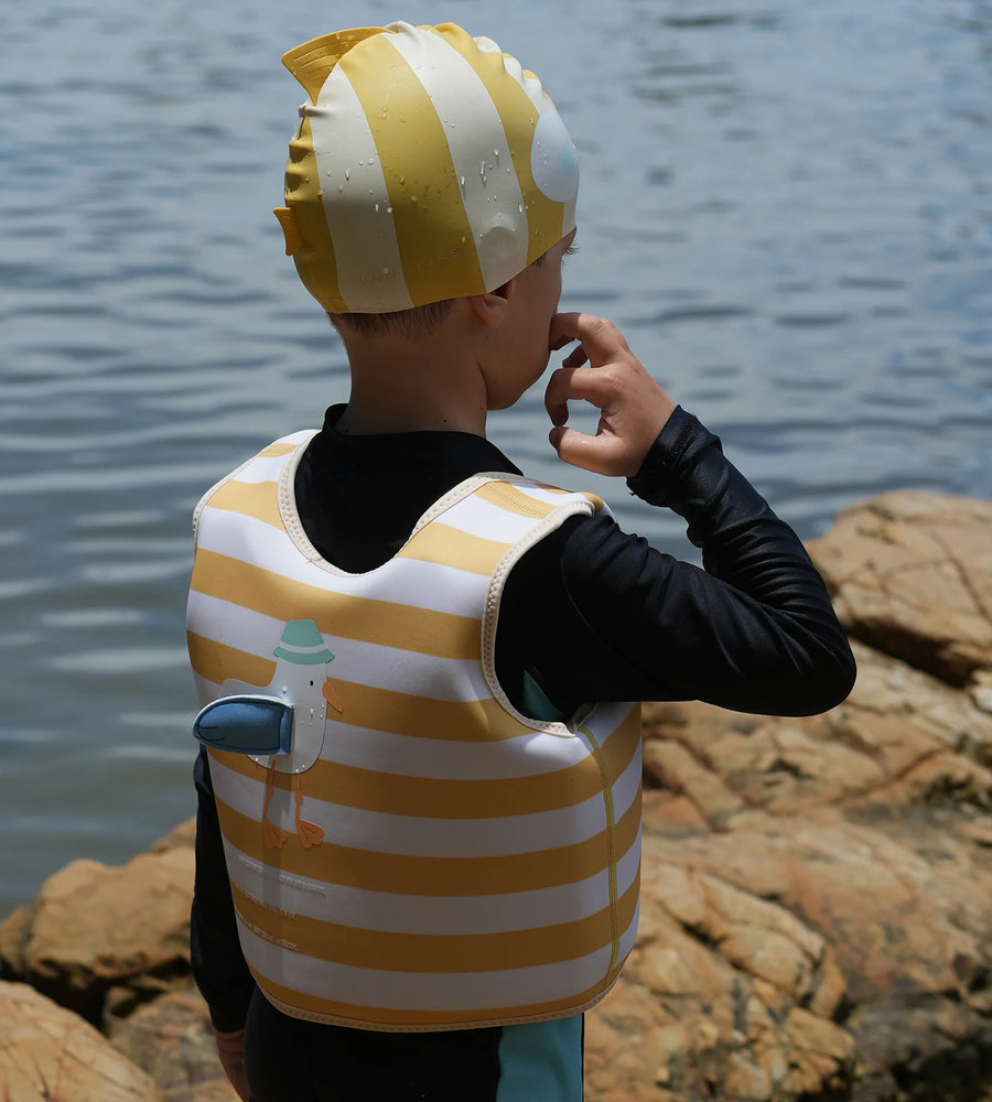 Child wearing a yellow and white striped swim cap and life jacket by a body of water.