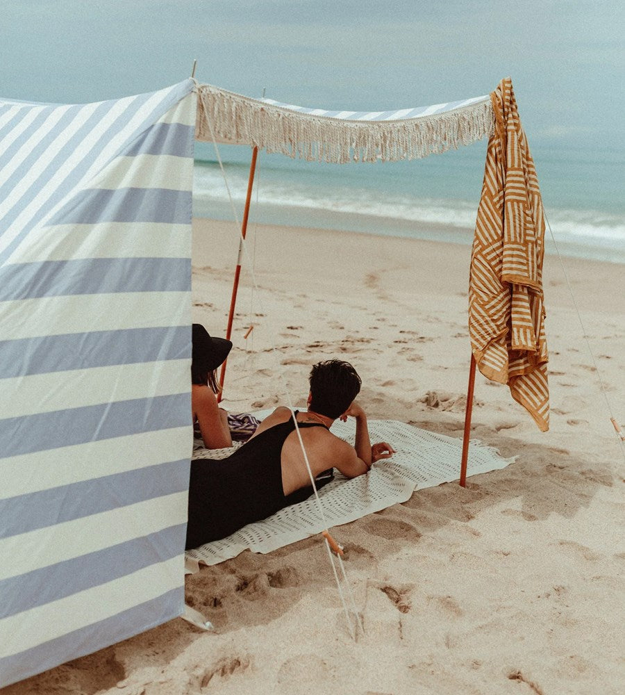 Two people sitting under a striped beach tent on a sandy beach with ocean view.