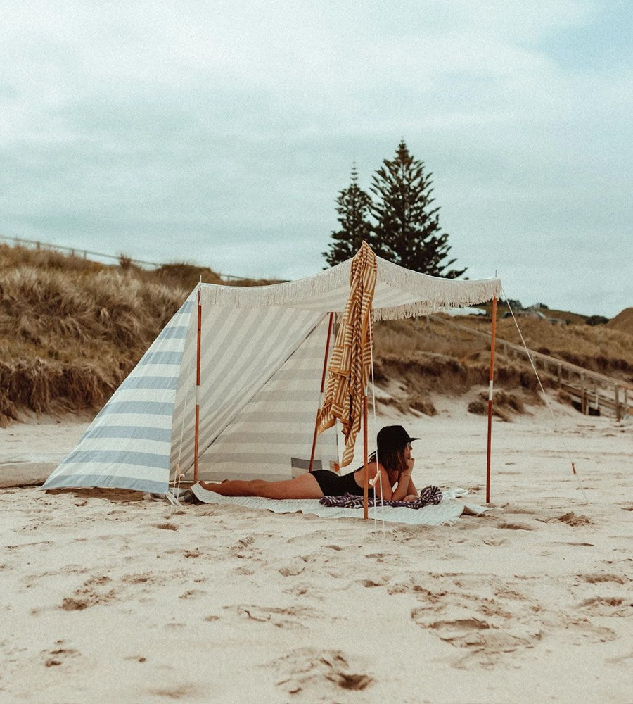 Person lying on a towel under a striped beach tent on a sandy beach with trees in the background.