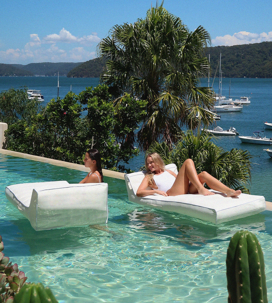 Two women lounging on inflatable pool floats by a pool with a scenic view of boats and trees.