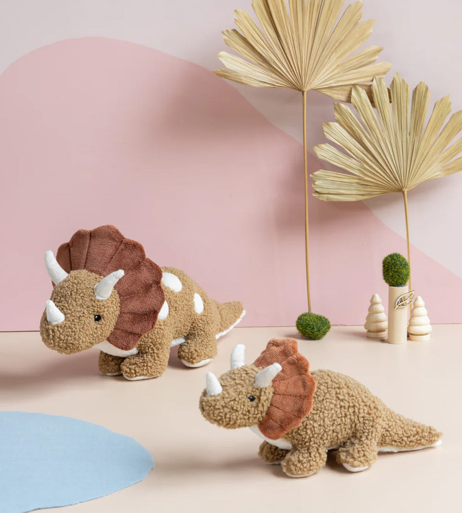 Two plush triceratops toys on a light pink surface with decorative plants in the background.