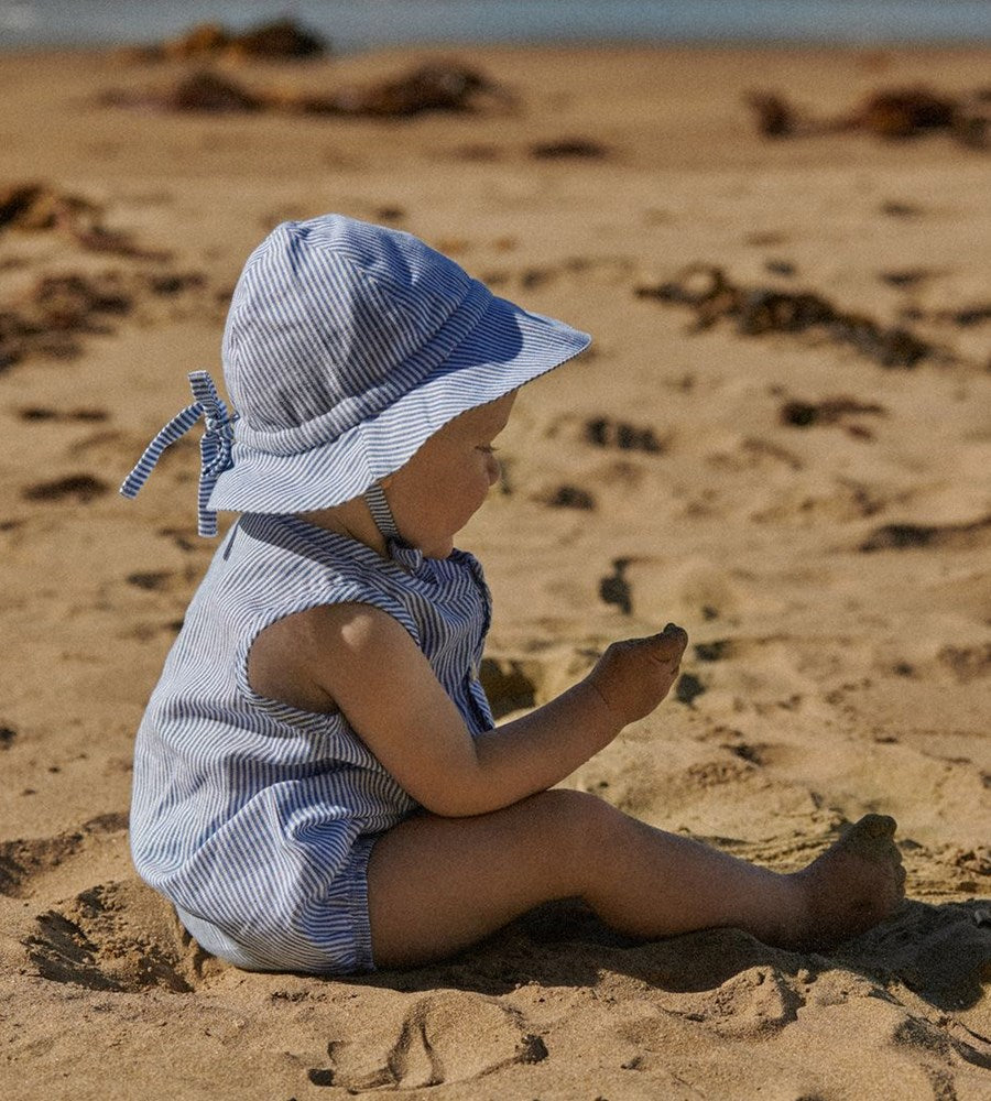 Child wearing a striped sun hat and romper sitting on a sandy beach.