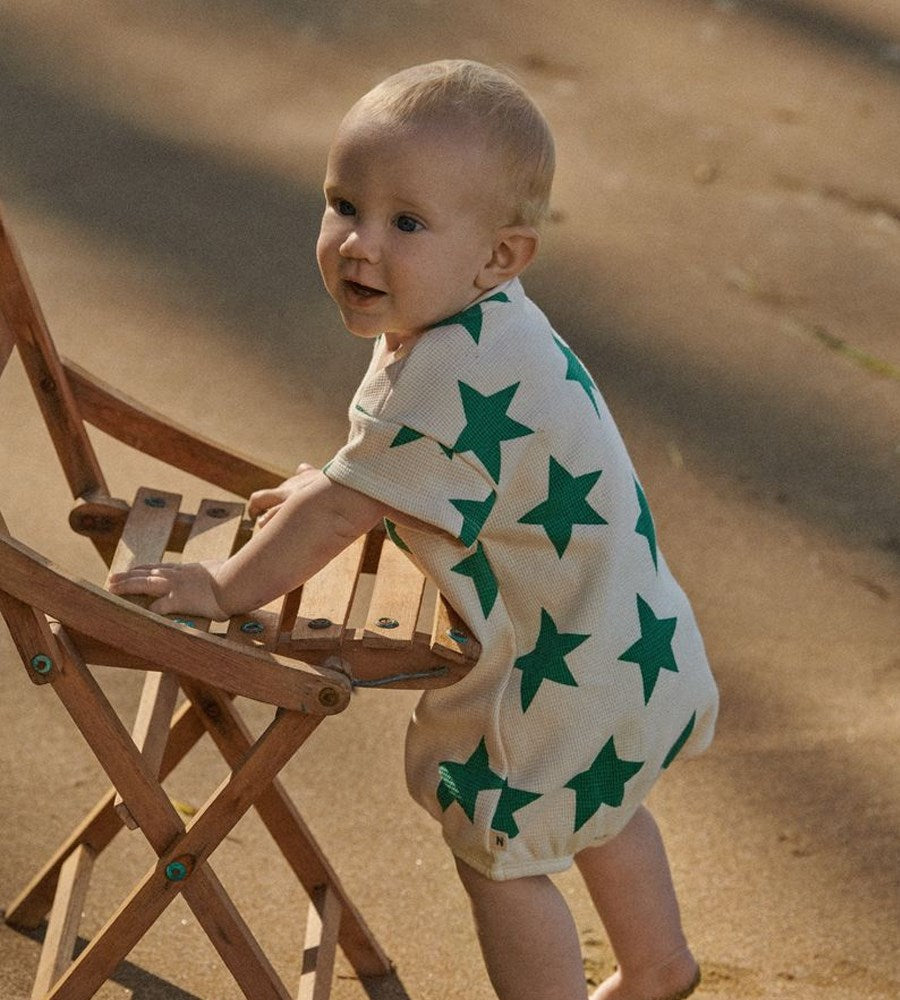 Baby wearing a white onesie with green star patterns, standing on a sandy surface.