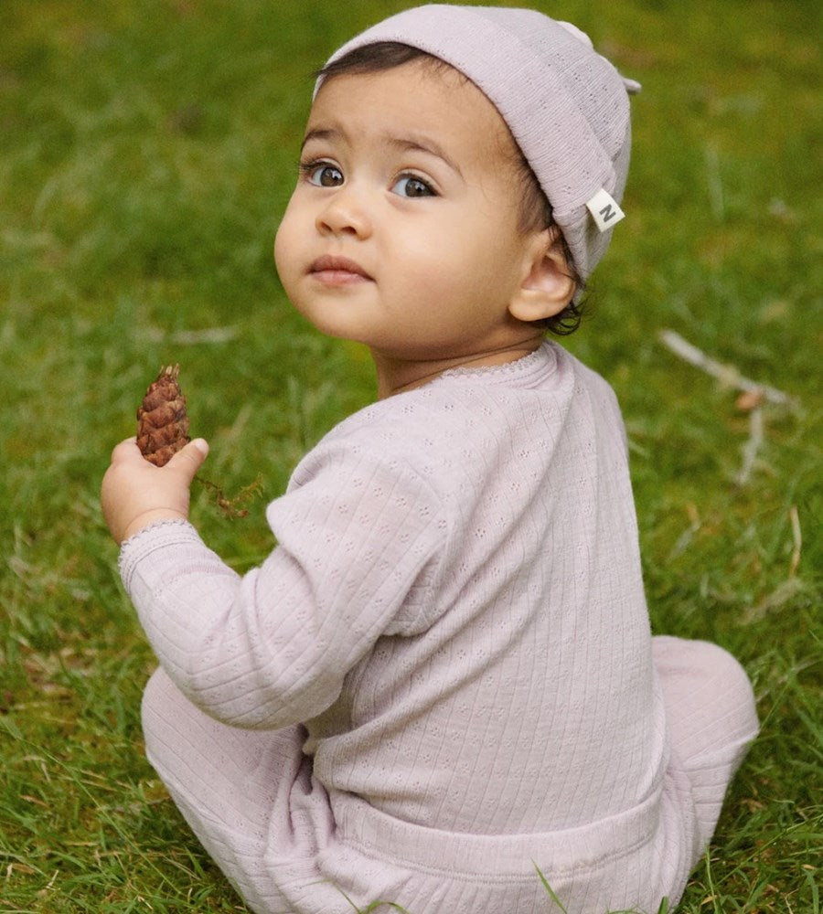 Baby in light purple outfit holding a pine cone in grass