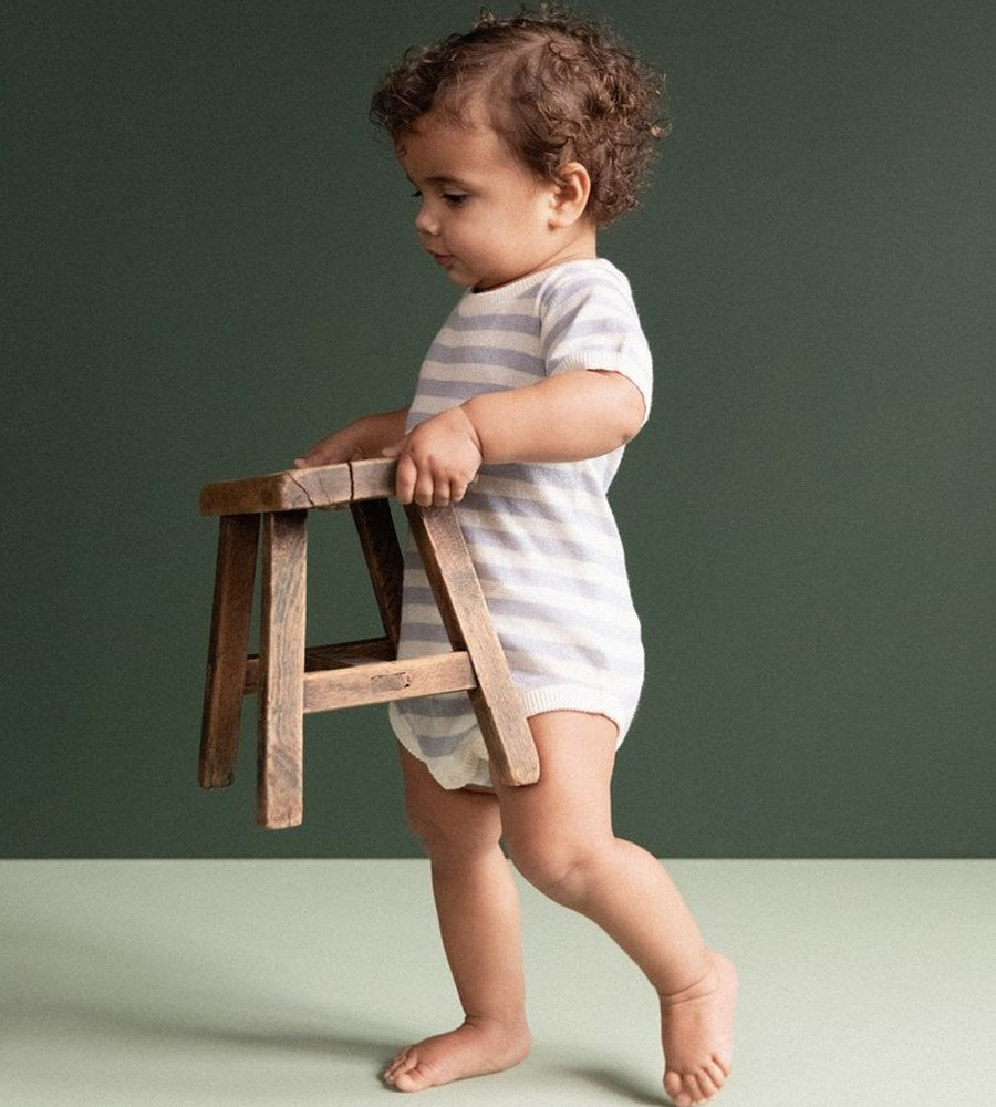 Child in a striped outfit standing next to a wooden stool against a green background