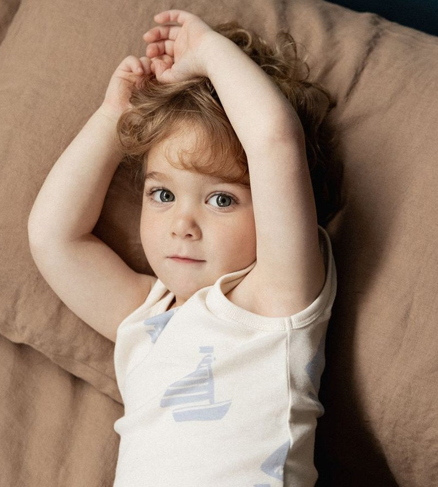 Child lying on a brown pillow with arms raised, wearing a white shirt with blue designs.