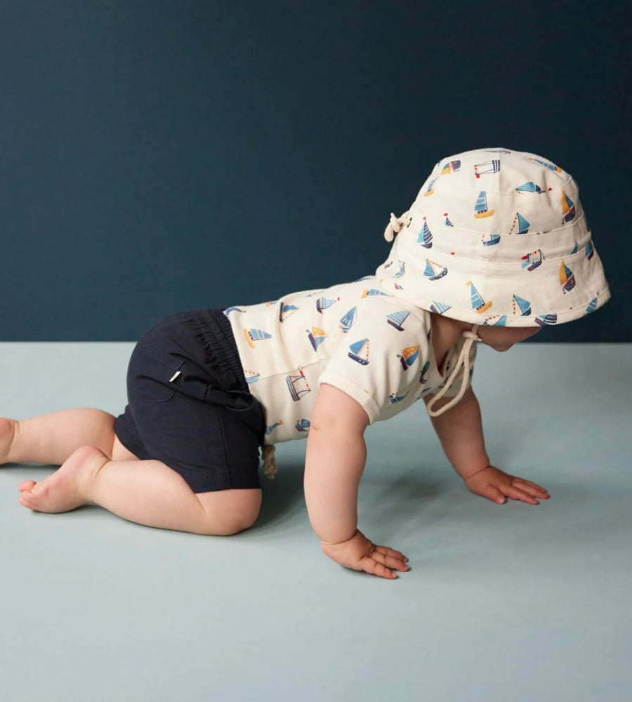 Baby wearing a sailboat-patterned sun hat and navy shorts on a blue background