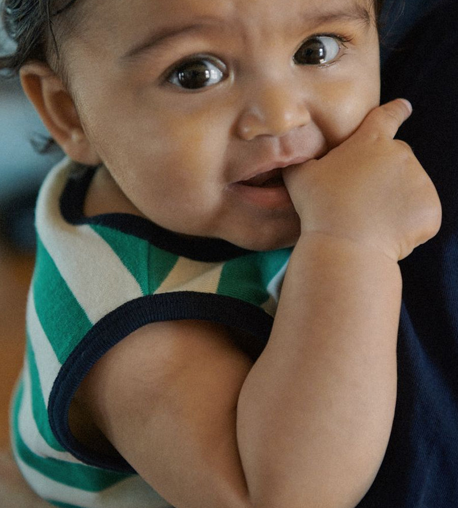 Baby wearing a green and white striped shirt with a dark collar, holding its hand to its mouth.