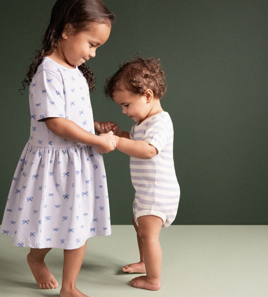 Two children standing on a plain background, one wearing a patterned dress and the other in a striped outfit.