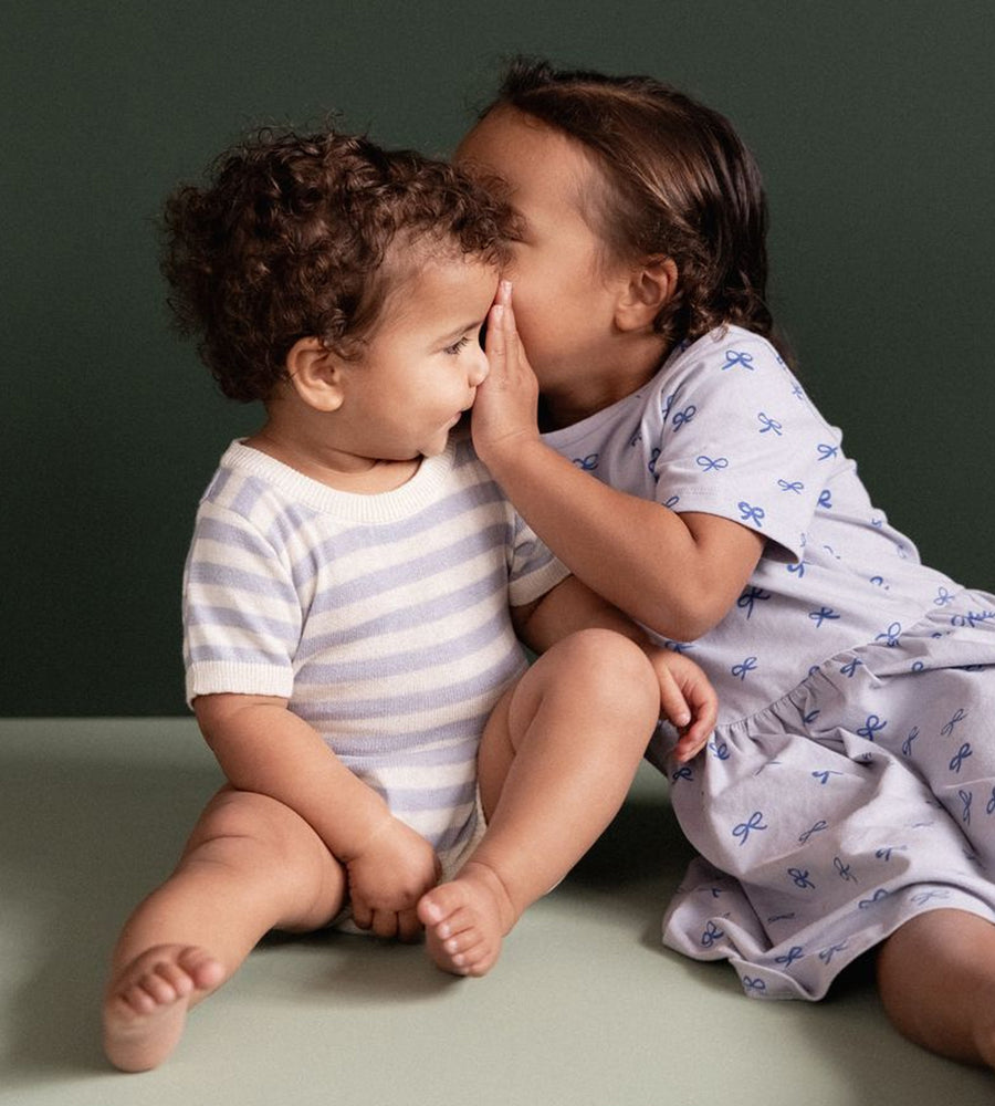 Two children sitting on a green surface, one whispering to the other against a dark green background.