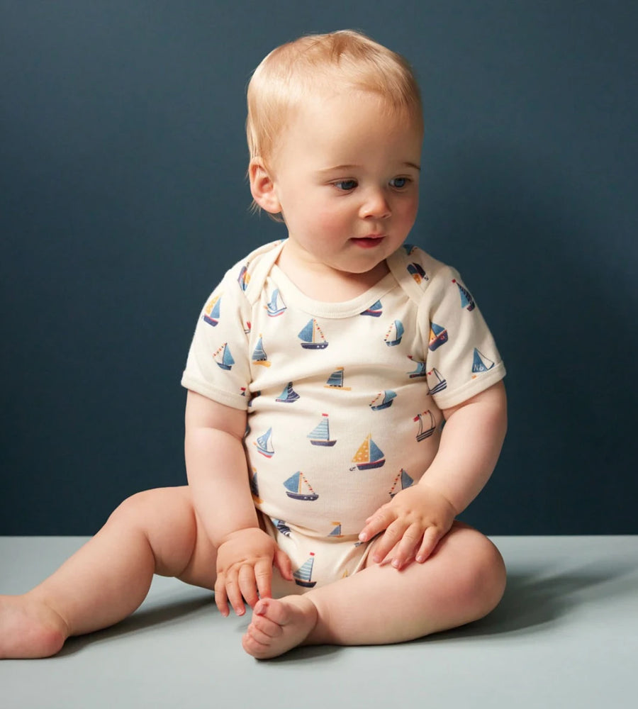 Baby sitting on a gray surface wearing a onesie with sailboat pattern against a dark blue background