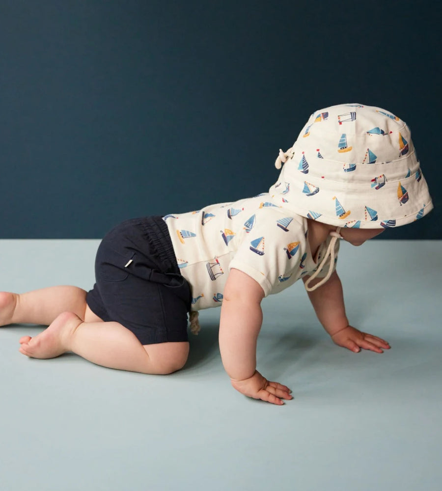 Child wearing a sailboat-patterned hat and navy shorts on a blue floor with dark blue wall background