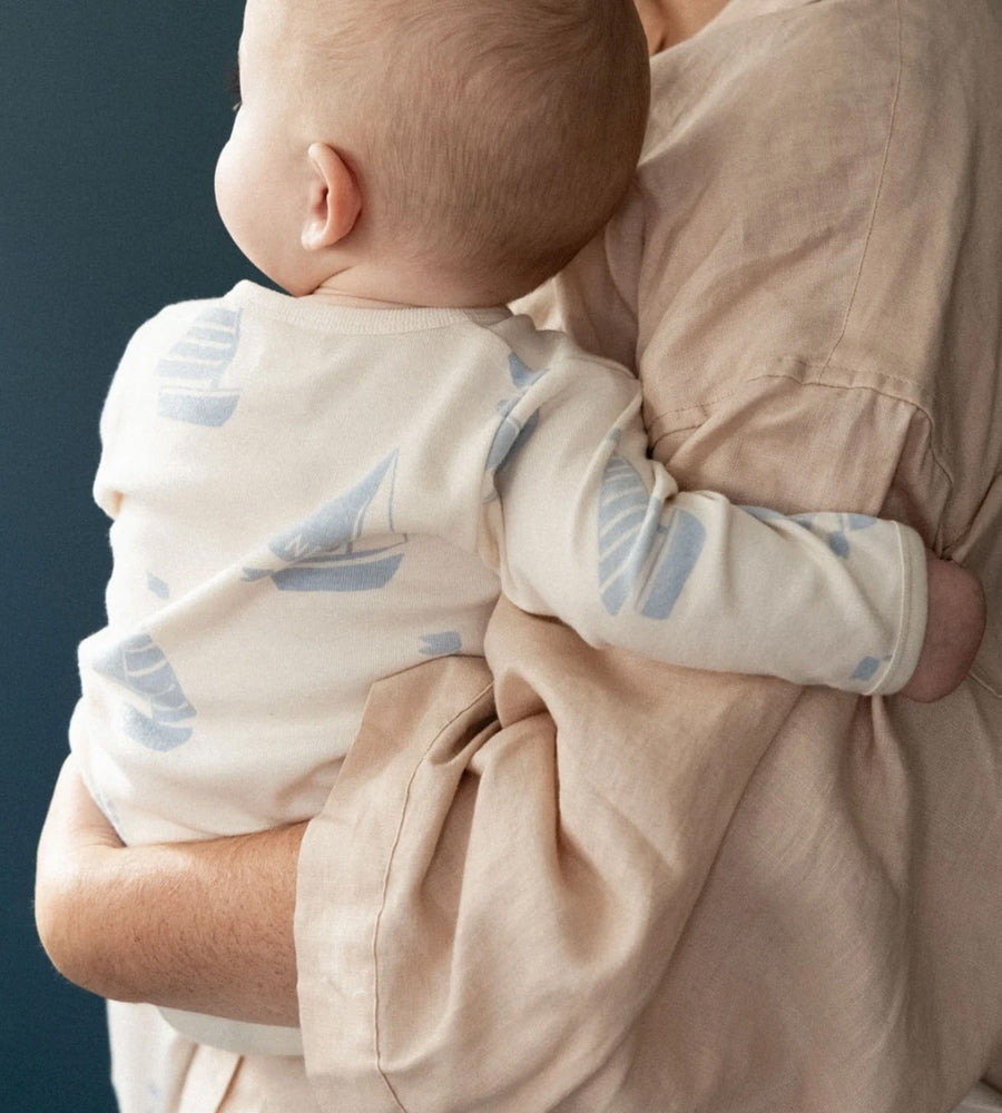 Baby wearing a white onesie with blue designs, held by an adult against a dark background
