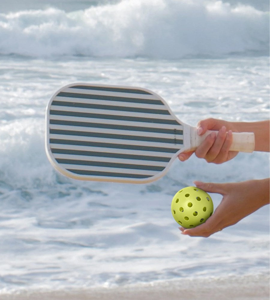 Hand holding a striped paddle and a yellow ball with ocean waves in the background