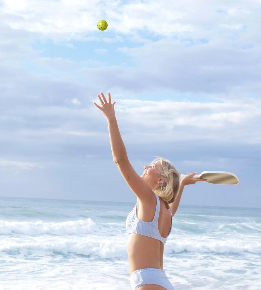Person playing beach cricket with a ball and bat, standing on a beach with ocean and sky in the background.
