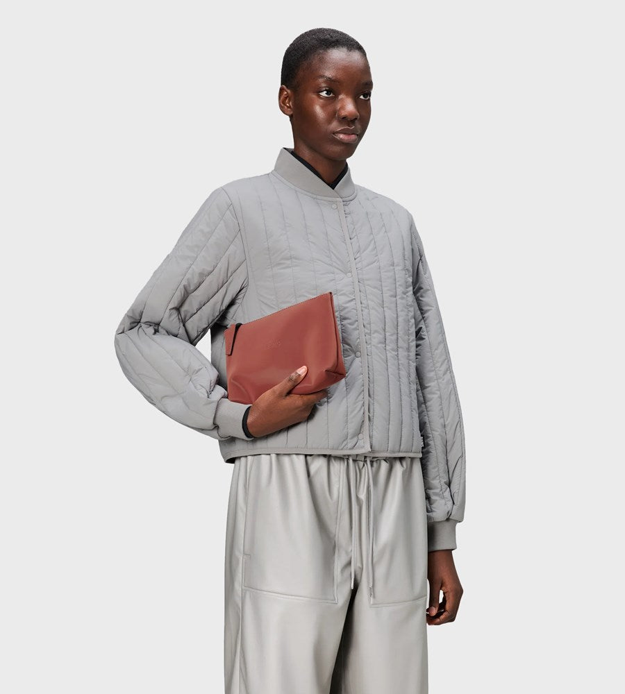 Person wearing a gray quilted jacket and beige pants holding a brown clutch against a light gray background