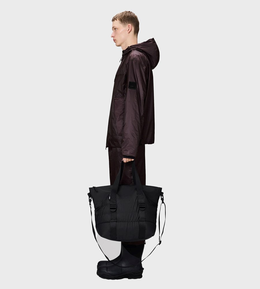 Person wearing a dark raincoat holding a black duffel bag on a white background