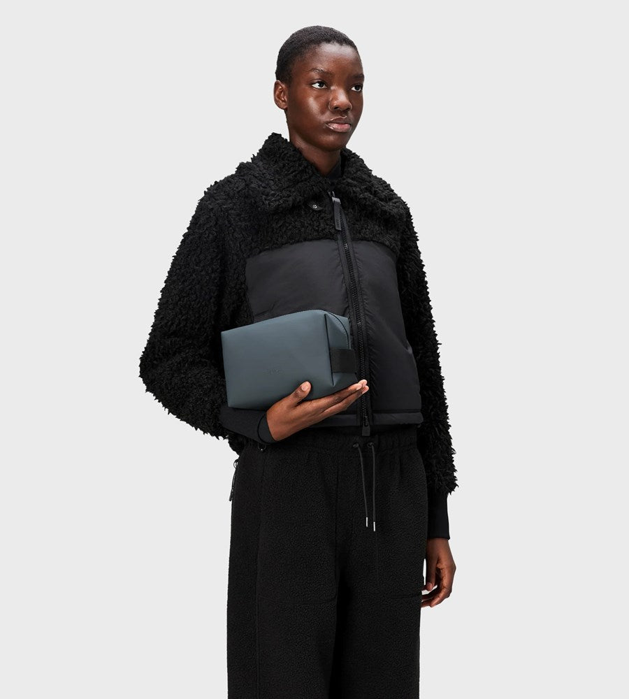 Person wearing a black shearling jacket holding a gray toiletry bag against a white background