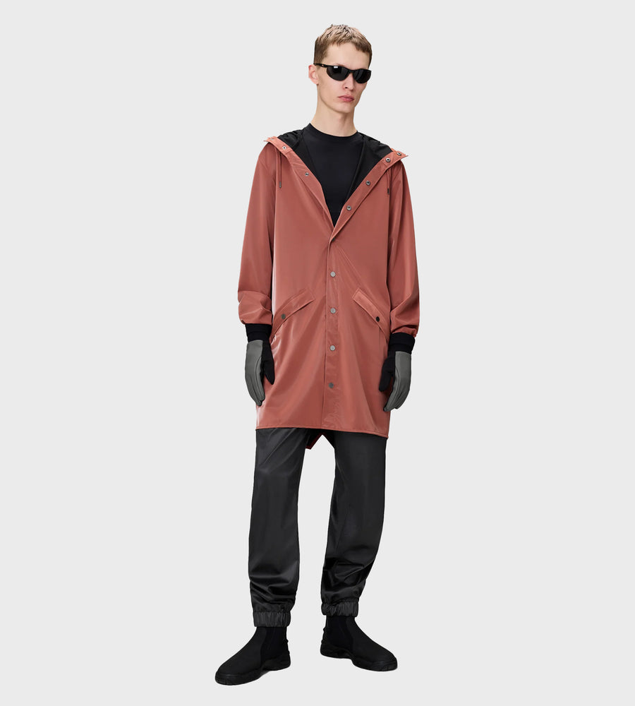 Person wearing a long rust-colored coat over a black outfit on a white background
