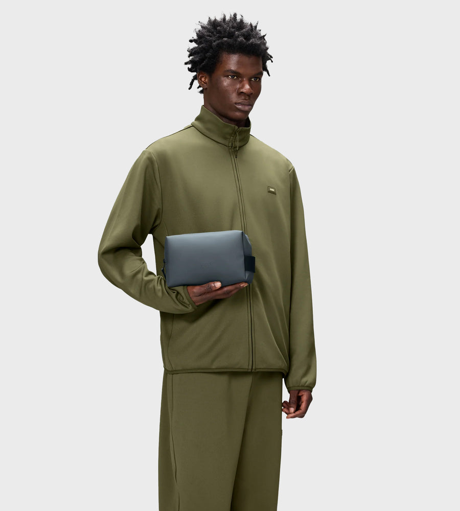 Person wearing a green tracksuit holding a gray toiletry bag on a white background