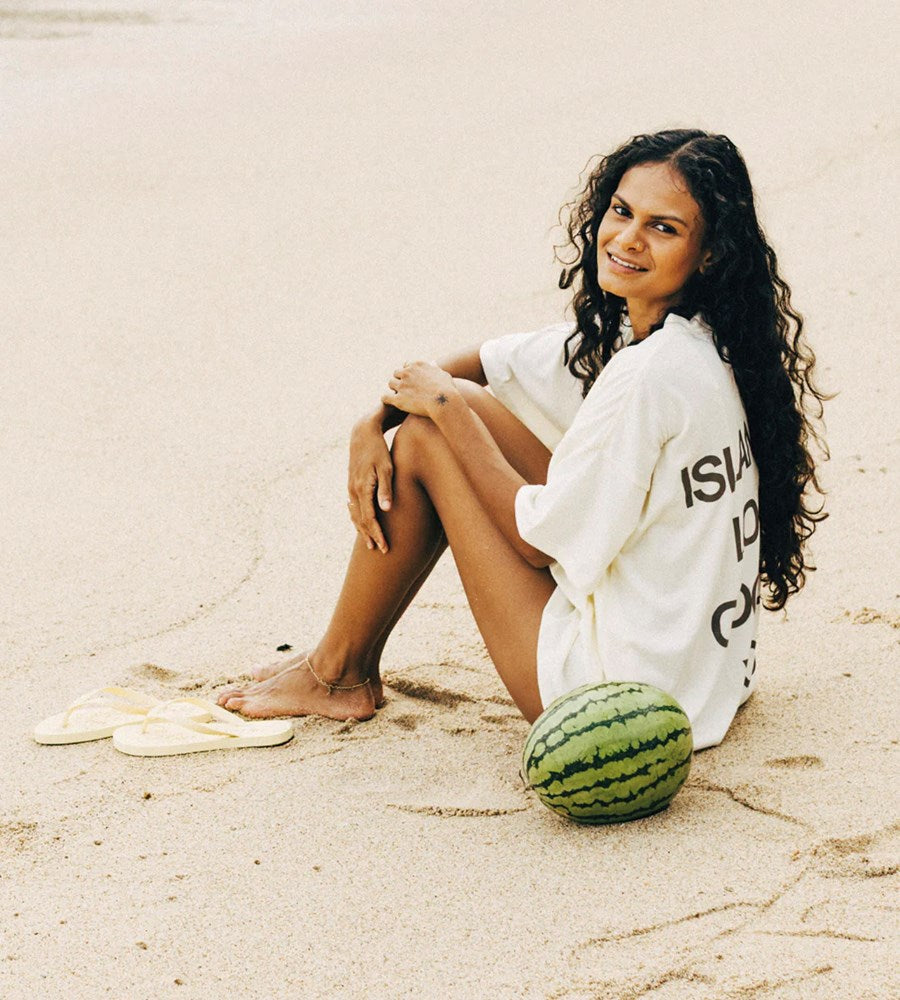 Woman sitting on sand with a watermelon, wearing a white outfit.
