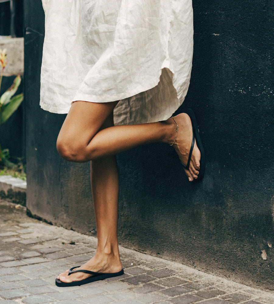 Person wearing a white outfit and black sandals against a dark wall.