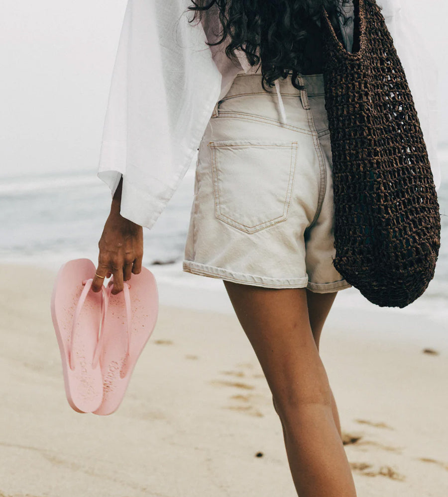 Person on a beach holding pink sandals and a woven bag
