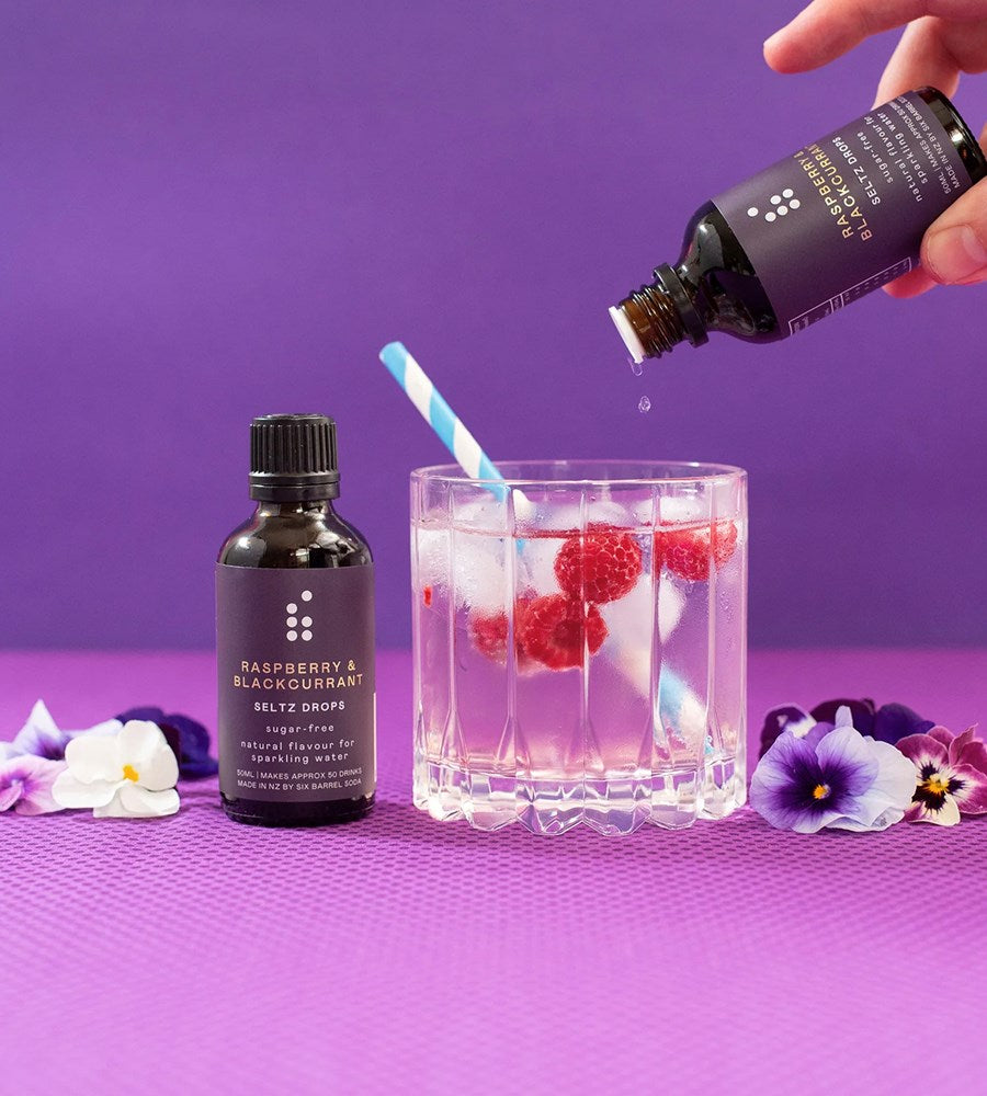 Bottle of raspberry and blackcurrant syrup being poured into a glass with ice and berries on a purple background.