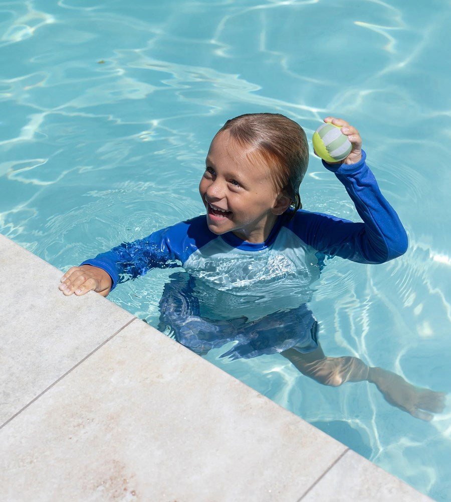 Child in a pool holding a ball, wearing a blue long-sleeve shirt.