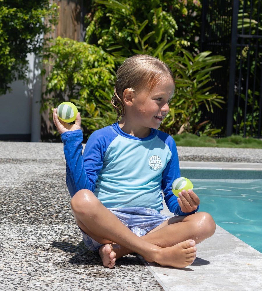 Child sitting by a pool holding two balls, wearing a blue and gray swimsuit.