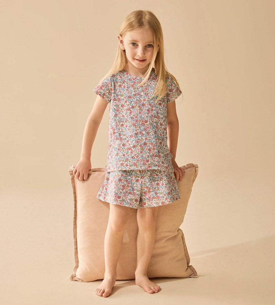 Young girl wearing a floral pajama set standing next to a beige pillow on a beige background