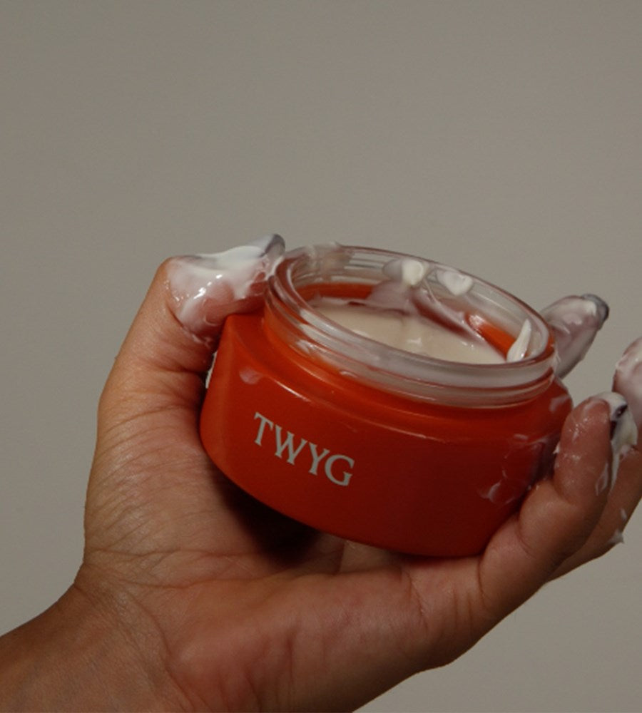 Red jar of TWYG cream held in a hand with a neutral background