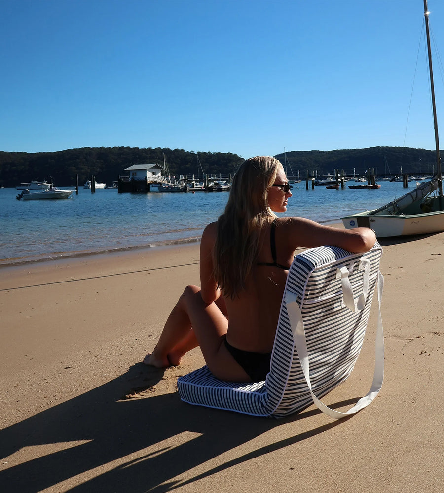 Woman sitting on a striped beach bag by the water with boats in the background
