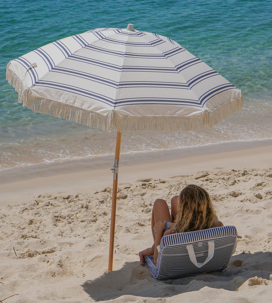 Woman sitting under a striped beach umbrella on a sandy beach with ocean in the background