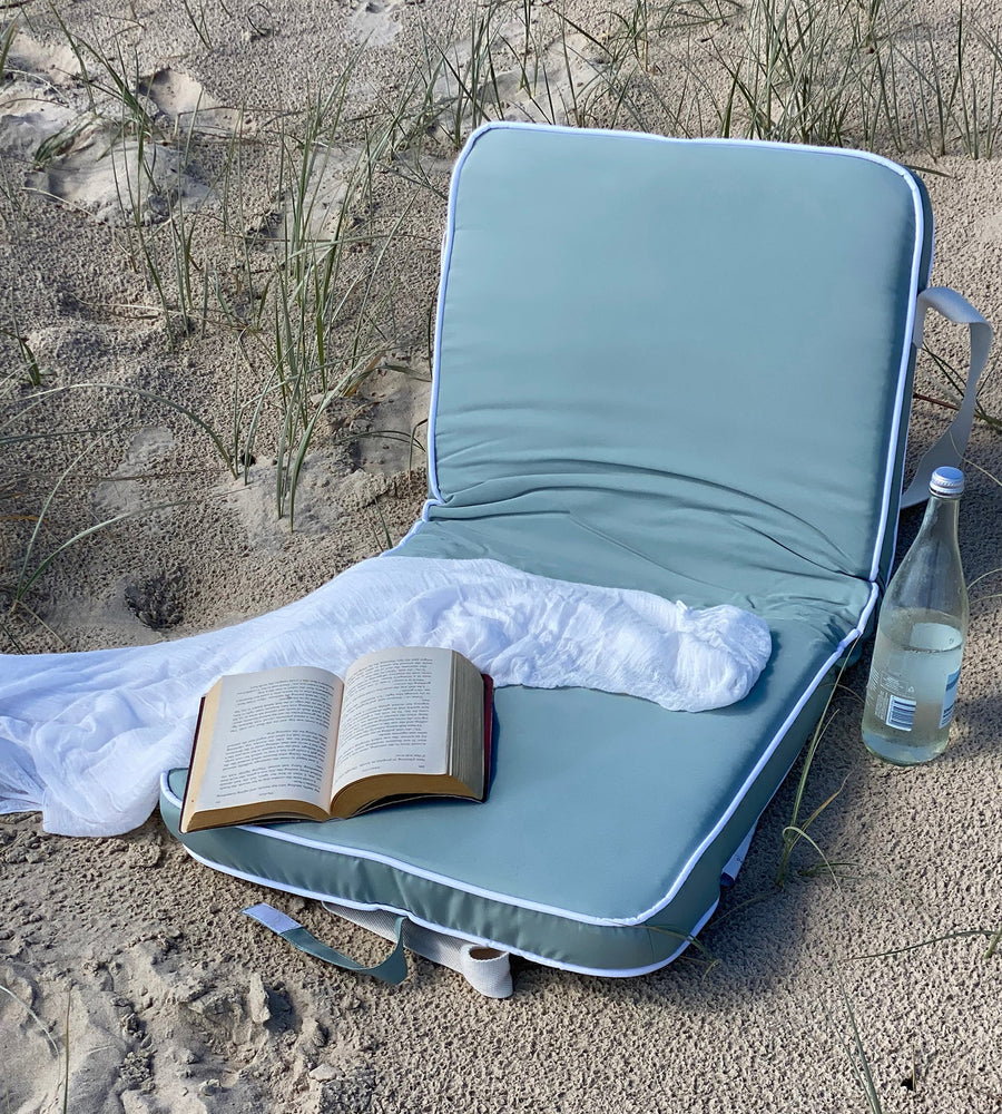 Light blue beach chair with an open book, white blanket, and bottle on sandy ground