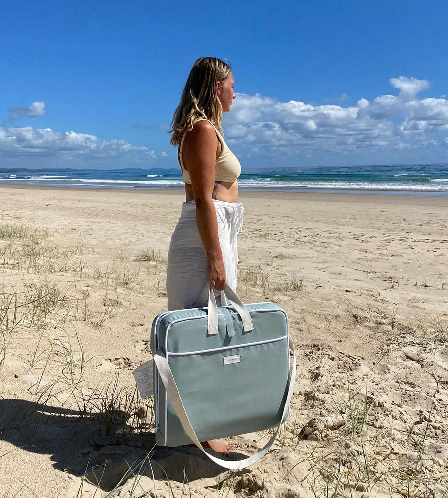 Woman holding a gray bag on a sandy beach with ocean and sky in the background
