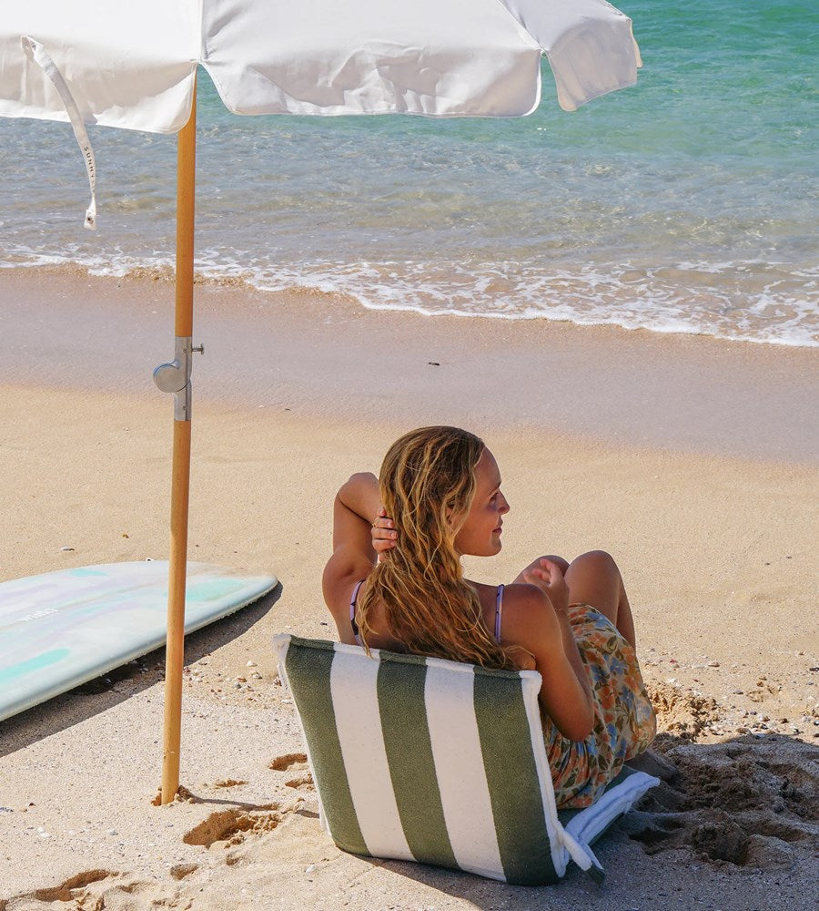 Woman sitting on a striped beach bag under an umbrella on a sandy beach with ocean in the background