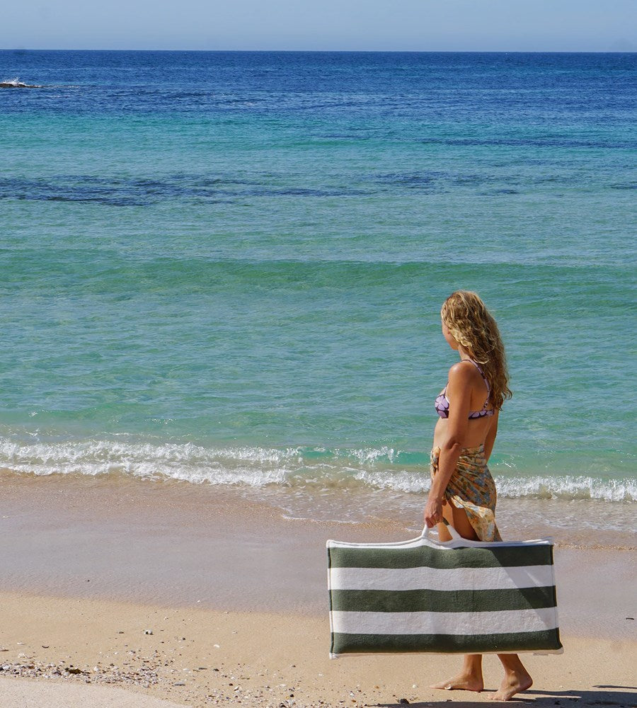 Woman holding a striped beach bag on a sandy beach with ocean in the background